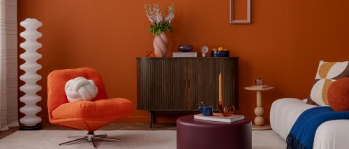 This retro-inspired living space, featuring a bold orange accent wall, plush white chairs, sculptural decor, and soft pastel accessories, perfectly captures the charm of new features in contemporary interior design. The playful yet sophisticated arrangement showcases how vintage elements can be refreshed with modern touches to create a vibrant and stylish home environment.
