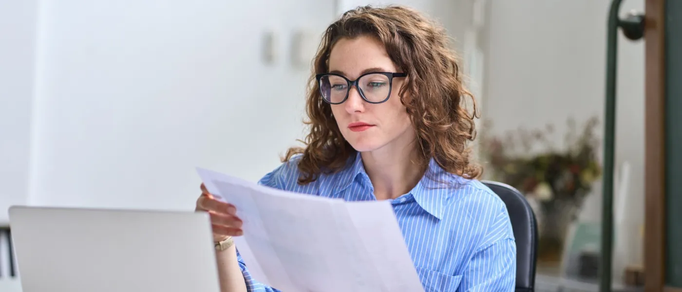 This image shows a young busy businesswoman managing accounting and financial documents while working on her laptop in a modern office. She appears focused on paperwork management and reviewing important data, reflecting the dedication and planning often required to pay rent between jobs and stay on top of expenses during transitional periods.