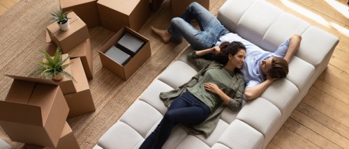 A couple sitting on a couch, surrounded by boxes, about to start settling into a new home.