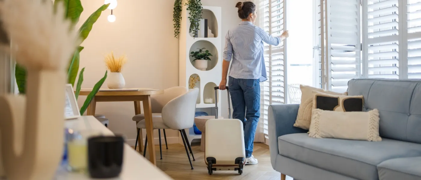 Woman with a suitcase closing window shutters in a bright, modern living room, illustrating vacation safety tips when leaving the apartment.