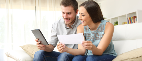 A smiling couple sits on a couch, using a tablet and holding a document to compare renters insurance policies.