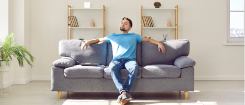 Relaxed man sitting on a grey sofa in his living room, enjoying fresh air after opening the windows, as a tip for spring home maintenance.